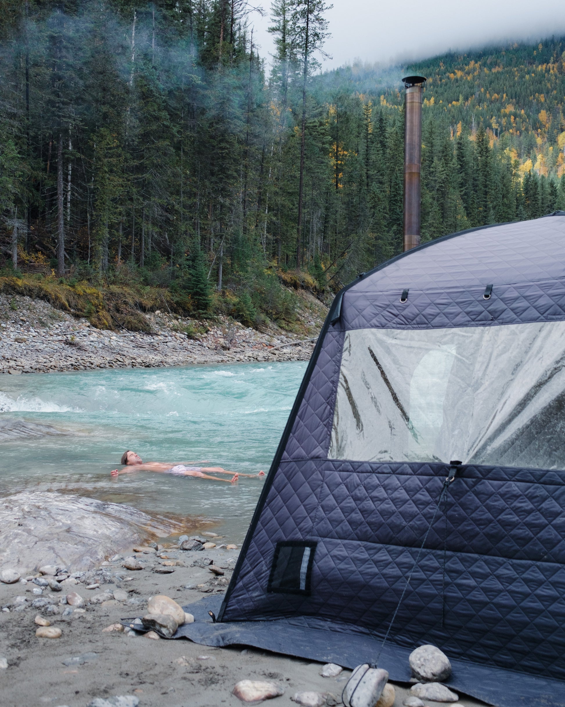 Person swimming in a river next to an outdoor sauna tent with a forest and mountains in the background