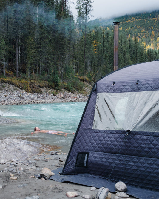 Person swimming in a river next to an outdoor sauna tent with a forest and mountains in the background