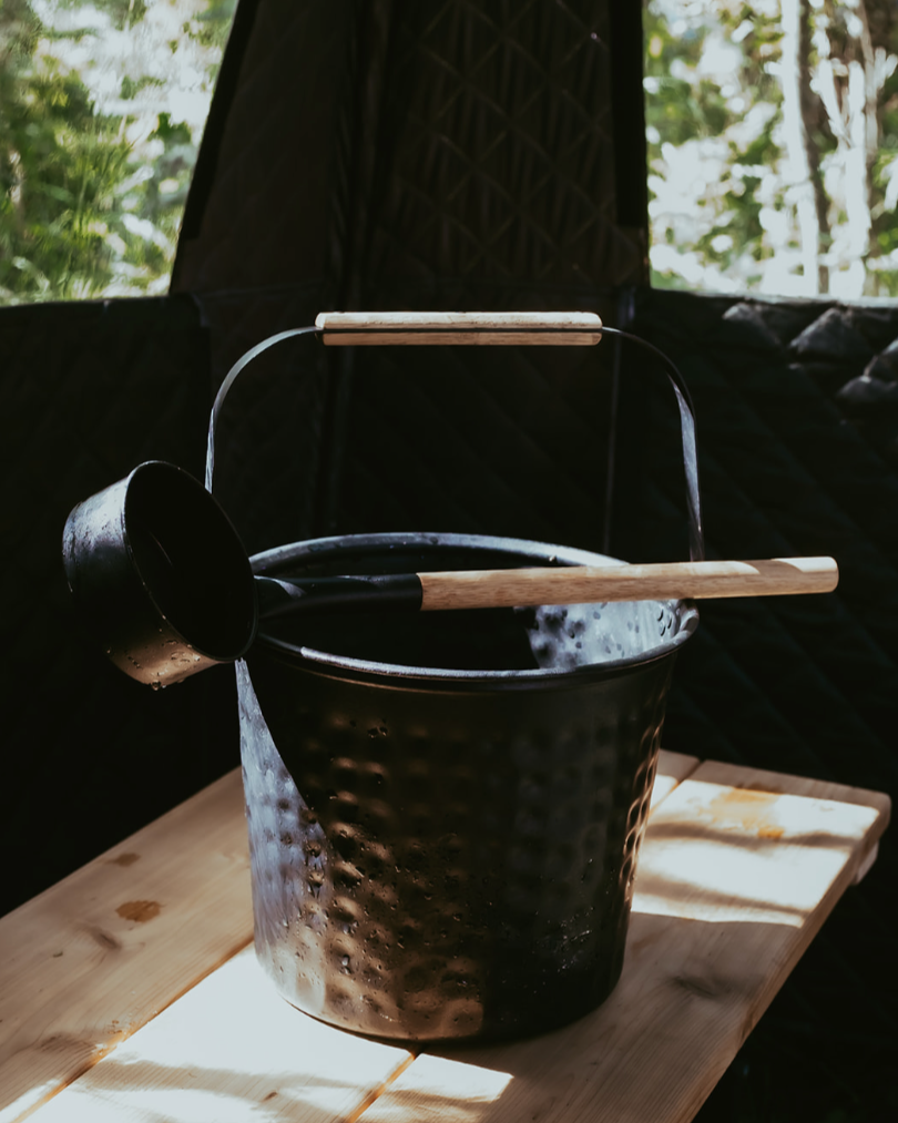 Black metal bucket with wooden handle on a wooden surface with blurred greenery in the background