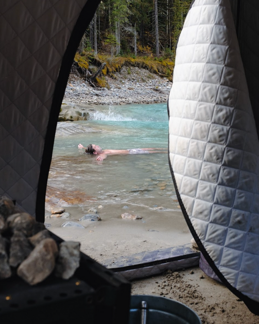 Inside view of a Thermaculture portable sauna tent with rocks and stove visible, overlooking a person cooling off in a mountain river.