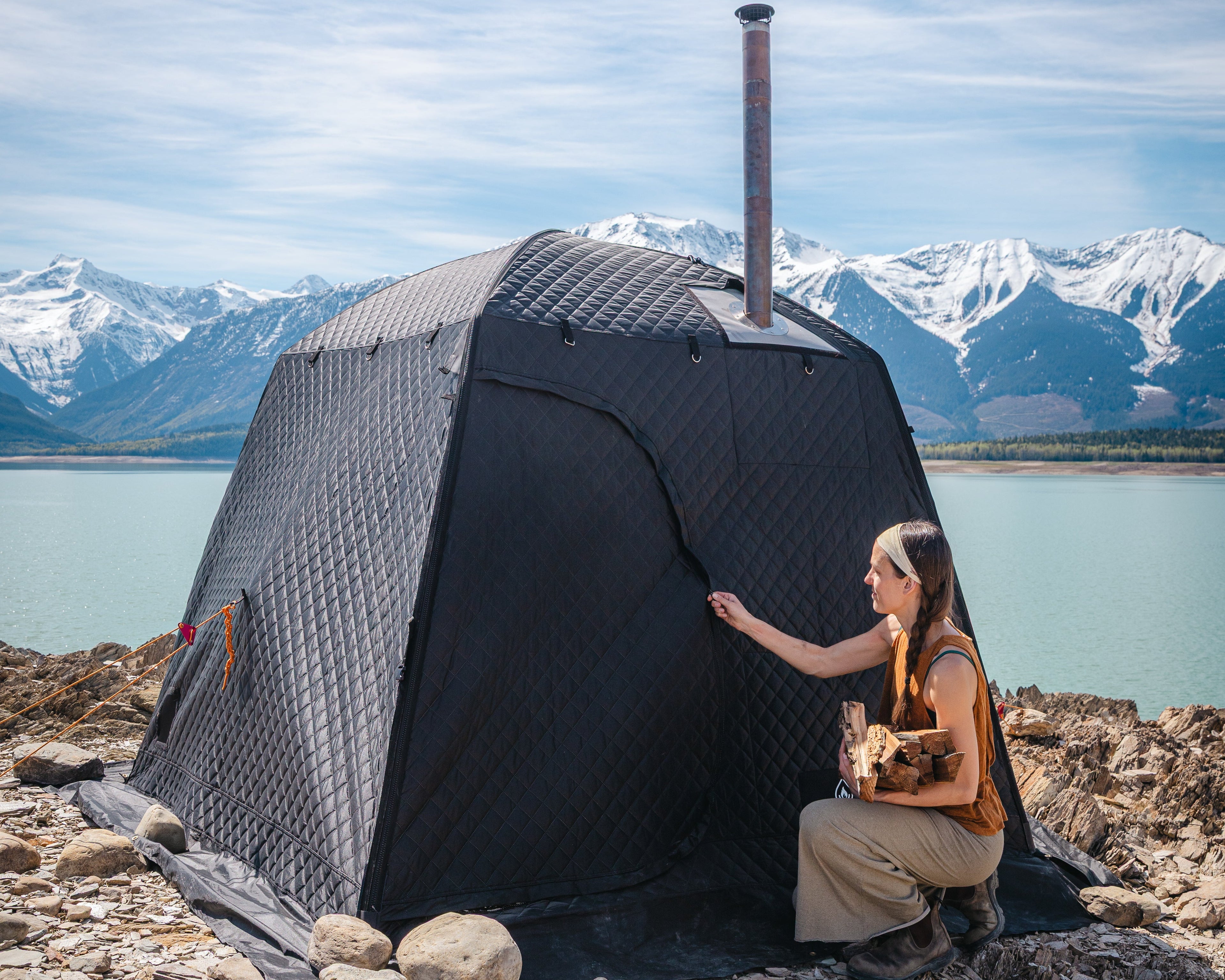 Person holding firewood sitting next to a large portable sauna tent on a rocky mountain landscape with mountains and water in the background
