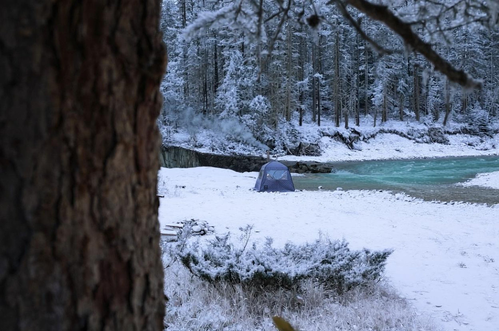 Person sitting in a Thermaculture sauna tent in a snowy forest with a river in the background